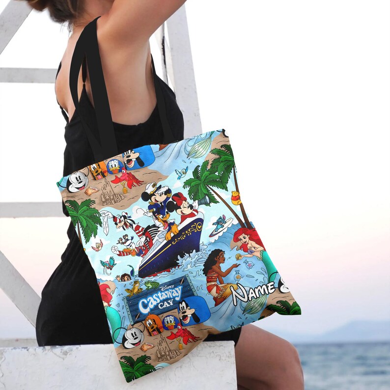 May include: A blue and white tote bag with a Disney Castaway Cay theme. The bag features Mickey Mouse, Minnie Mouse, Donald Duck, Goofy, and other Disney characters. The bag also has the text "Disney Castaway Cay" and "Name".