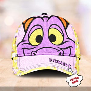 May include: A purple and yellow baseball cap with a cartoon dragon face on the front. The dragon has large yellow eyes and a purple snout. The cap has the word "FIGMENT" written on the brim. The cap is customizable with a name.
