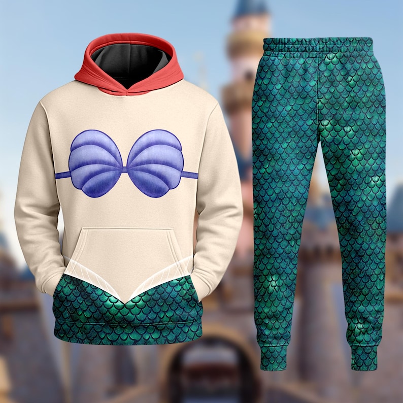 Op de afbeelding: Een beige hoodie en bijpassende broekenset. De hoodie heeft een blauw schelpenbh-ontwerp en een rood gevoerde capuchon. De broek heeft een groen schubbenpatroon. De set is ontworpen om op een zeemeermin te lijken.