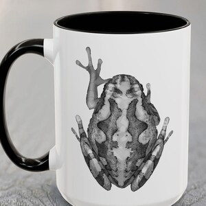 May include: White ceramic coffee mug with a black rim and handle. The mug features a black and white illustration of a frog on the front.