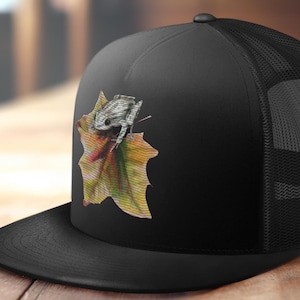 May include: Black trucker hat with a black mesh back. The front of the hat features a colorful embroidered image of a frog sitting on a leaf.