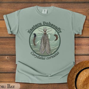 May include: A bay-colored t-shirt featuring a circular graphic with an illustration of an Eastern Dobsonfly, a larva, and the text "Eastern Dobsonfly Corydalus cornutus". The shirt is made of soft material.