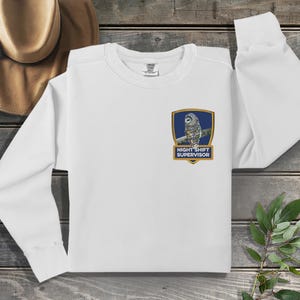 May include: White long-sleeve sweatshirt with a blue and gold patch featuring an owl and the text "Night Shift Supervisor".