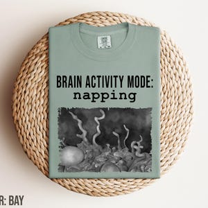 Peut inclure: Un t-shirt vert sauge avec une image en noir et blanc d'un cerveau et le texte "Brain Activity Mode: napping".