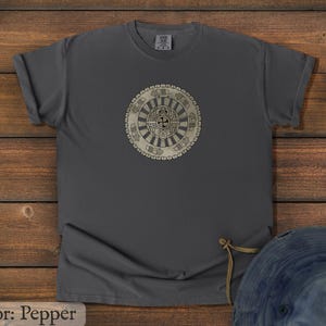 May include: Pepper-colored t-shirt with a circular, beige graphic in the center. The graphic features a detailed, symmetrical design with a central cross-like shape and surrounding patterns. The shirt is made of a soft material and has rolled-up sleeves.