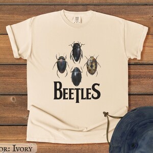 May include: Ivory-colored t-shirt featuring a graphic of four beetles in various colors above the word "BEETLES" in a bold, black font. The beetles are in shades of black, brown, and tan. The shirt is made of a soft material.
