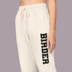 Könnte beinhalten: Cremefarbene Jogginghose mit Kordelzug in der Taille. Das Wort "BIRDER" ist vertikal in fetten, schwarzen Buchstaben auf dem linken Bein aufgedruckt. Die Hose ist aus einem weichen Material.