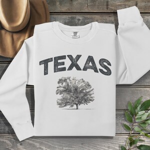 Könnte beinhalten: Weißer Langarm-Pullover mit dem Wort "TEXAS" in Schwarz auf der Brust gedruckt. Unter dem Text befindet sich eine Schwarzweißzeichnung eines Baumes.