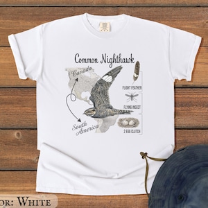 May include: White t-shirt featuring a detailed illustration of a Common Nighthawk, with text labels for flight feather, flying insect, and egg clutch. The design includes a map of Canada and South America. The shirt is white.