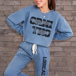 Può includere: Un completo composto da felpa e pantaloni della tuta blu chiaro. La felpa presenta la scritta "ORNI TWO" in un carattere nero stilizzato. I pantaloni della tuta hanno la parola "LOGICAL" stampata verticalmente sulla gamba sinistra. Il completo è realizzato in materiale morbido.