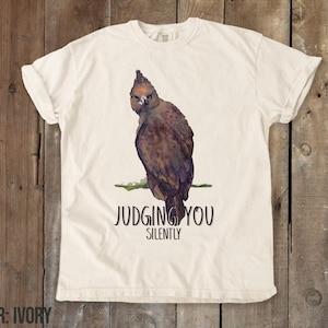 Puede incluir: Camiseta de color marfil con una ilustración de un pájaro marrón y negro y el texto "Judging You Silently".