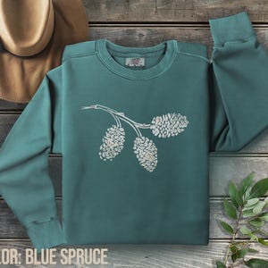 Könnte beinhalten: Ein tealblaues Sweatshirt mit einem weißen Kiefernzweig-Design. Das Sweatshirt hat einen Rundhalsausschnitt und lange Ärmel. Der Text "COLOR: BLUE SPRUCE" befindet sich am unteren Rand des Bildes. Ein beiger Hut ist im Hintergrund.
