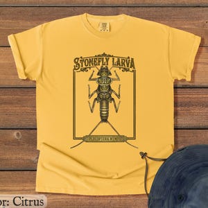 May include: Citrus-colored t-shirt featuring a detailed illustration of a stonefly larva within a rectangular frame. The text "STONEFLY LARVA" is arched above the insect, with "PLECOPTERA NYMPH" below. The shirt is made of a soft material.