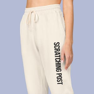 May include: Cream-colored sweatpants with a drawstring waist. The text "SCRATCHING POST" is printed vertically in black on the left leg. The pants are made of a soft material.