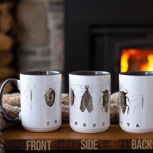 Peut inclure: Trois tasses en céramique blanche avec des bords et des poignées noirs. Chaque tasse présente une illustration d'insecte différente et le mot "INSECT" imprimé sur le côté. Les tasses sont disposées en rangée sur une surface en bois.