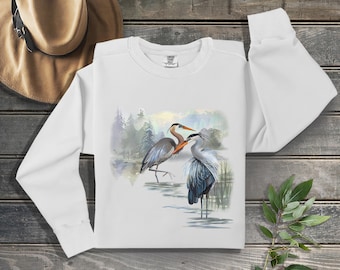 May include: White long-sleeved sweatshirt with a watercolour illustration of two herons in a marsh. The birds have grey and blue feathers, with orange beaks and necks. The background shows a forest and a colourful sky.