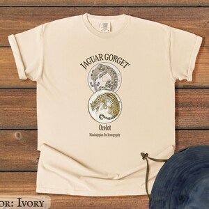 May include: Ivory-colored t-shirt featuring a graphic design with the text "JAGUAR GORGET" above two circular illustrations of a jaguar and an ocelot. The text "Ocelot" and "Mississippian Era iconography" are below the illustrations. The shirt is made of a soft material.