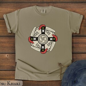 May include: Khaki-colored t-shirt featuring a circular design with a central spiral and stylized bird-like elements in red, black, and white. The shirt is made of a soft material and has a classic crew neck.