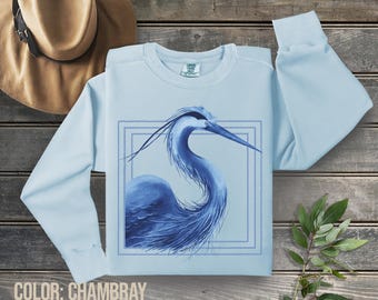 May include: A light blue sweatshirt featuring a blue heron illustration within a pale blue frame. The heron is facing right, with a long neck and beak. The text "COLOR: CHAMBRAY" is at the bottom of the image.