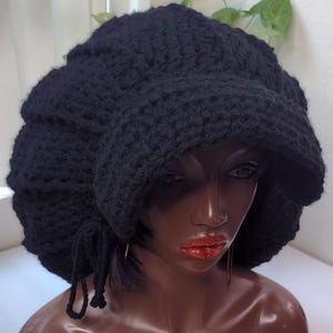 May include: A solid black crocheted newsboy cap featuring a thick, textured yarn construction with a prominent brim and a decorative side tie detail. The hat has a rounded, voluminous crown and is displayed on a mannequin head to show its shape.