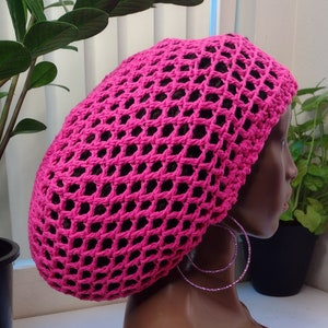 May include: A bright pink, open-weave crochet hat. The hat is displayed on a brown mannequin head, with a brown crocheted collar. The mannequin is wearing hoop earrings. The background includes potted plants.
