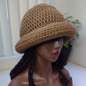 May include: A brown crochet bucket hat with a textured, open-stitch crown and a solid, wide brim. The hat is displayed on a mannequin head featuring dark brown skin, long dark twisted hair, and thin gold hoop earrings, wearing a brown crochet top.
