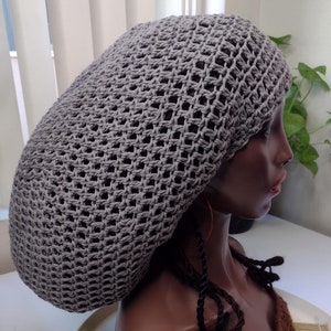 May include: A gray crocheted hat with an open weave design. The hat is displayed on a mannequin head with dark brown skin. The mannequin also has dark brown braids and a brown crocheted top.