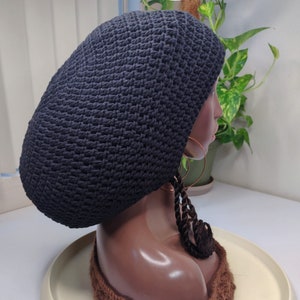 Peut inclure: Un bonnet noir en crochet avec de longues tresses qui pendent dans le dos. Le bonnet est sur une tête de mannequin avec une écharpe en crochet marron autour du cou.