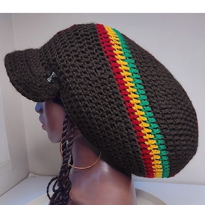 Peut inclure: Un bonnet en crochet marron avec une visière noire et une bande rouge, jaune et verte. Le bonnet est porté sur un mannequin aux cheveux bruns.