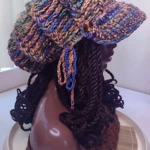 K&ouml;nnte beinhalten: Eine grob gestrickte Schieberm&uuml;tze mit einem mehrfarbigen Garnmuster in Violett, Blau, Gr&uuml;n und Gold. Die M&uuml;tze verf&uuml;gt &uuml;ber einen kurzen Schirm, einen dekorativen kupferfarbenen Spiralknopf an der Seite und zwei herabh&auml;ngende geflochtene Quasten.