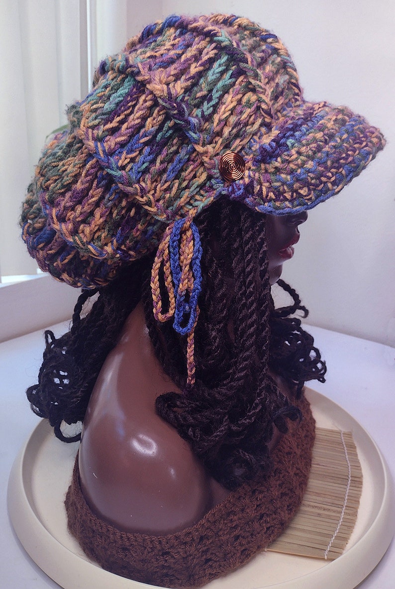 K&ouml;nnte beinhalten: Eine grob gestrickte Schieberm&uuml;tze mit einem mehrfarbigen Garnmuster in Violett, Blau, Gr&uuml;n und Gold. Die M&uuml;tze verf&uuml;gt &uuml;ber einen kurzen Schirm, einen dekorativen kupferfarbenen Spiralknopf an der Seite und zwei herabh&auml;ngende geflochtene Quasten.
