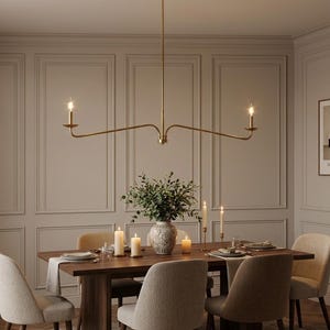 Op de afbeelding: Een eethoek met een donkerhouten eettafel en een goudkleurige kroonluchter. De kroonluchter heeft drie brandende lampen. Beige gestoffeerde stoelen staan rond de tafel, die gedekt is voor een maaltijd. Een vaas met groen staat in het midden.