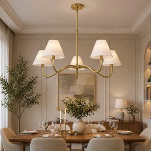 May include: A gold-toned chandelier with five white lampshades hangs above a wooden dining table set for a meal. The room features neutral tones, a large painting, and a vase of greenery. The chandelier's design is elegant and adds a touch of sophistication.