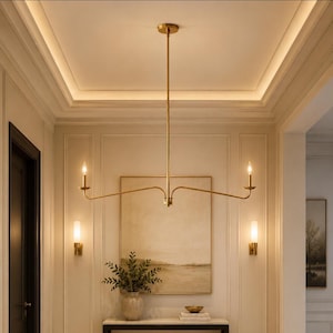 Pode incluir: Um lustre de metal em tom dourado com uma longa haste vertical e dois braços horizontais que se estendem para fora, cada um terminando em uma luminária vertical estilo vela. A peça possui um acabamento liso, polido e simétrico.