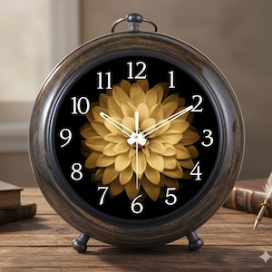 May include: A round, antique-style clock with a black face featuring white numerals and a golden flower design. The clock has a bronze-colored metal frame and stands on two small feet. The clock also has a handle at the top.