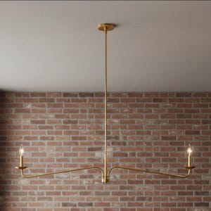 Puede incluir: Una lámpara de araña dorada de dos luces, con una varilla vertical central y brazos horizontales. Cada brazo tiene una sola lámpara con una bombilla en forma de vela. La lámpara está montada en un techo blanco contra una pared de ladrillo.