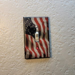 Könnte beinhalten: Ein Lichtschalter-Abdeckung mit einem gedruckten Bild der amerikanischen Flagge. Die Flagge ist rot, weiß und blau mit einem verwitterten Look. Der Schalter ist weiß und hat einen schwarzen Umriss.