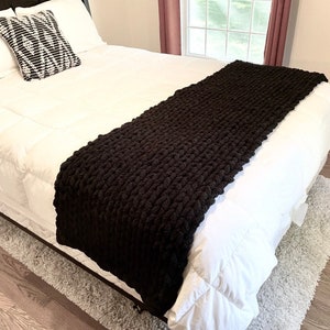 Könnte beinhalten: Eine schwarze, dicke Strickdecke, die über einem weißen Bett mit einem schwarz-weißen Kissen drapiert ist. Die Decke ist aus dickem Garn gefertigt und hat eine strukturierte Optik.