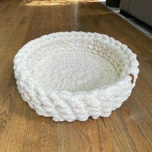 May include: A round, off-white pet bed made of thick, chunky knit yarn. The bed has a low profile with a raised edge and a small, brown leather tag. The texture of the yarn is visible, creating a cozy appearance.