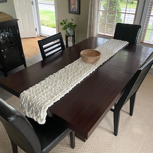 May include: A white knitted table runner with a chunky texture sits on a dark brown wooden table with black chairs. A wooden bowl sits on the table runner.