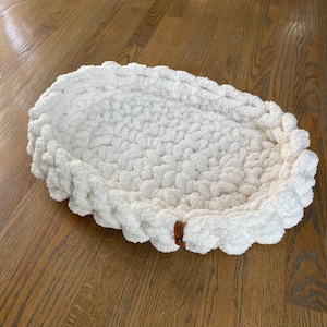 May include: A white, oval-shaped pet bed made of chunky, textured yarn. The bed has a raised, scalloped edge and a small, brown leather tag. The bed is designed for small pets.