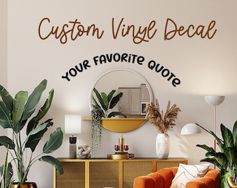 Puede incluir: Un interior de sala de estar con una calcomanía de vinilo personalizada que dice "Custom Vinyl Decal Your Favorite Quote". La calcomanía está en una pared blanca sobre un gabinete de oro y mimbre. Hay un gran sillón naranja, un otomano blanco y una alfombra con estampado en blanco y negro.