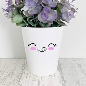 May include: A white ceramic plant pot with a cute face design, holding faux purple and green eucalyptus leaves. The pot has a smiling face with pink cheeks and a tongue sticking out. The pot is sitting on a white and gray wooden surface.