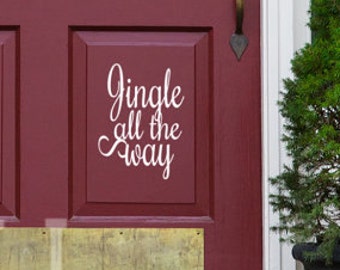 Può includere: Una porta d'ingresso rossa con un adesivo in vinile bianco che recita "Jingle all the way". La porta ha una piastra di protezione in ottone ed è fiancheggiata da due alberi sempreverdi in vaso.