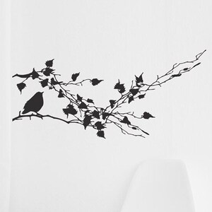 May include: Black silhouette wall decal of a bird perched on a branch with leaves. The decal is on a white wall with a white chair and a woven basket in the foreground.