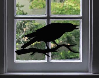 May include: A black crow silhouette decal on a white window frame. The crow is perched on a branch and is looking to the right. The window is behind the crow and shows a view of green trees.