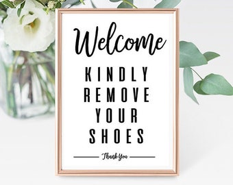 May include: A printable sign with a rose gold frame that says "Welcome Kindly Remove Your Shoes - Thank You". The sign is sitting on a white surface with flowers in the background.