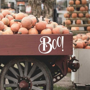 May include: A red wagon filled with orange pumpkins with the word "Boo!" written in white on the side. The wagon is parked in front of a pile of pumpkins.