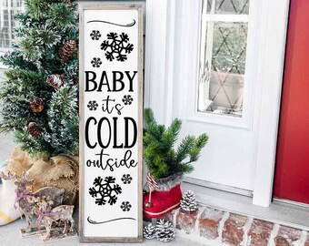以下が含まれることがあります： 雪の結晶のデザインが施された、「Baby it's cold outside」という黒い文字が書かれた白い木製の看板。看板は白いドアに寄りかかり、隣には赤いドアがある。看板の両側には、小さな人工のクリスマスツリーが2本ある。