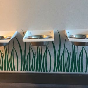 Puede incluir: Tres cuencos blancos para mascotas montados en la pared con cuencos de acero inoxidable. Los cuencos est&aacute;n montados sobre un dise&ntilde;o de hierba verde.